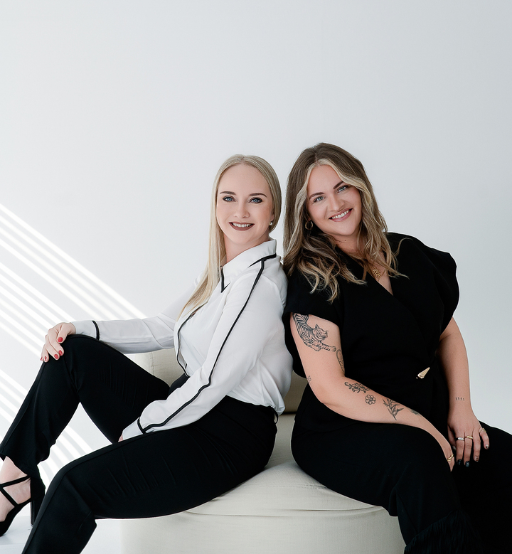 Two individuals seated closely on a cushioned bench in a bright minimalist room, one with a smartphone and the other with tattoos visible.