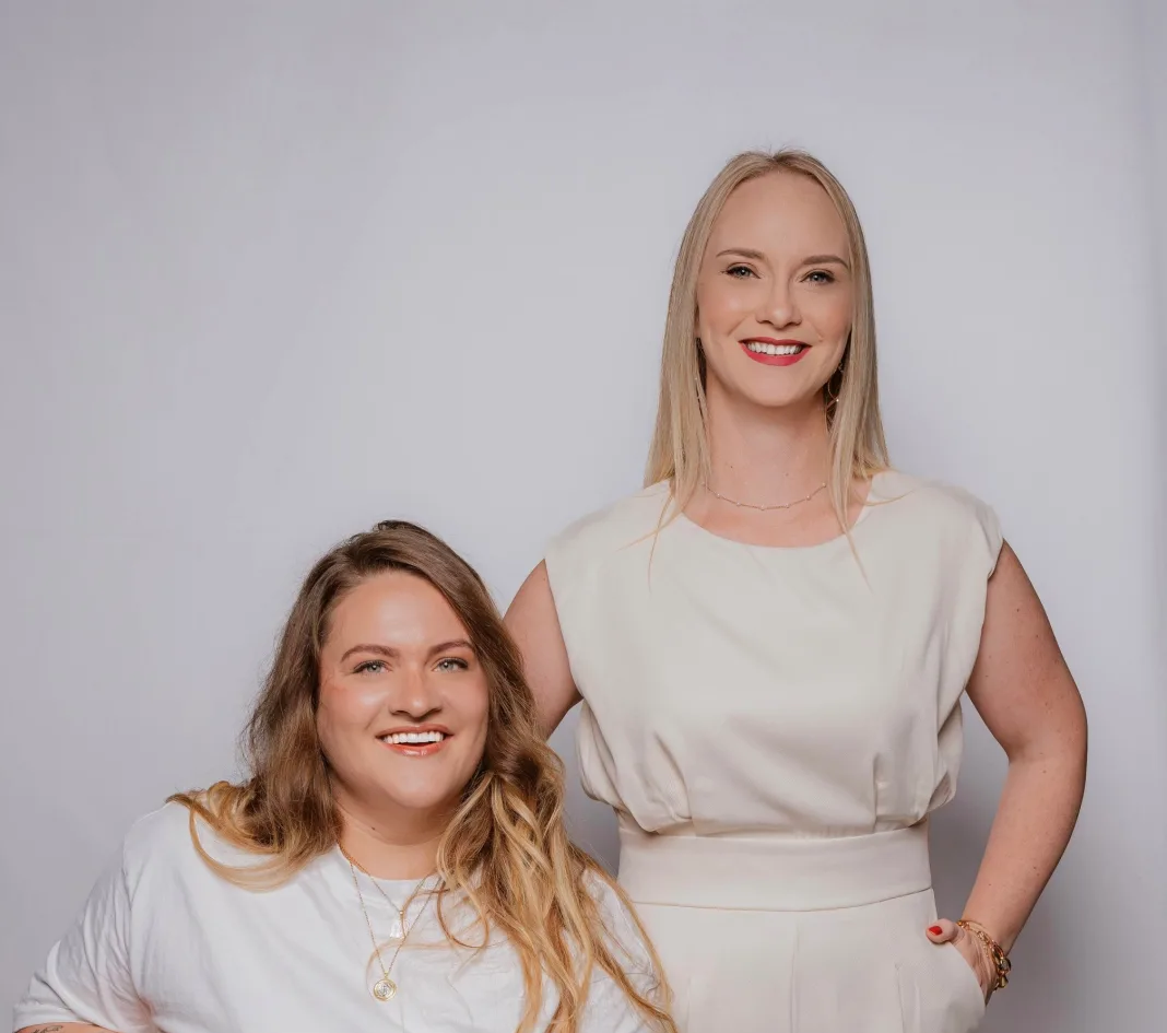Two smiling individuals posing together, one seated in a white shirt with layered necklaces and the other standing in a cream belted outfit.