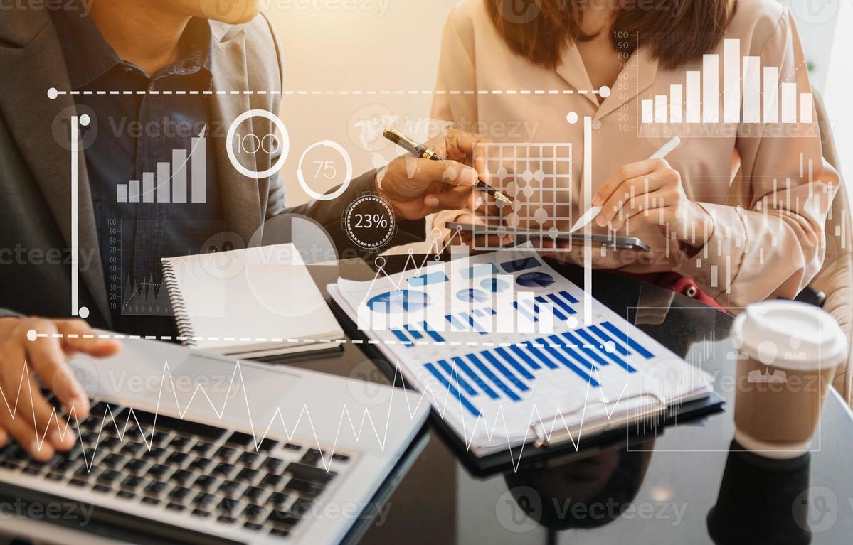 Two people at desk reviewing charts with a laptop, tablet, coffee, and overlaid graphs showing