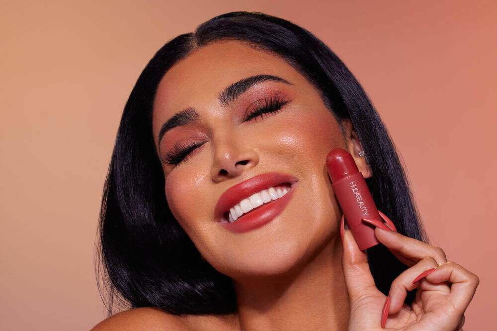 Close-up of woman holding Huda Beauty lipstick near face.