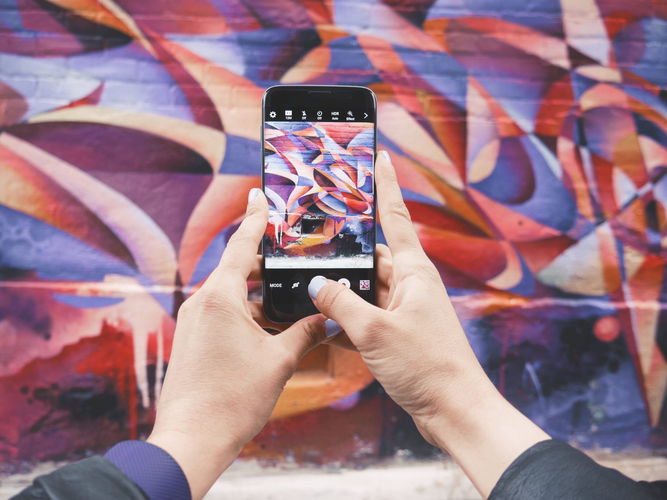 Services 2 Close‑up of hands holding smartphone photographing colourful abstract mural on brick wall.