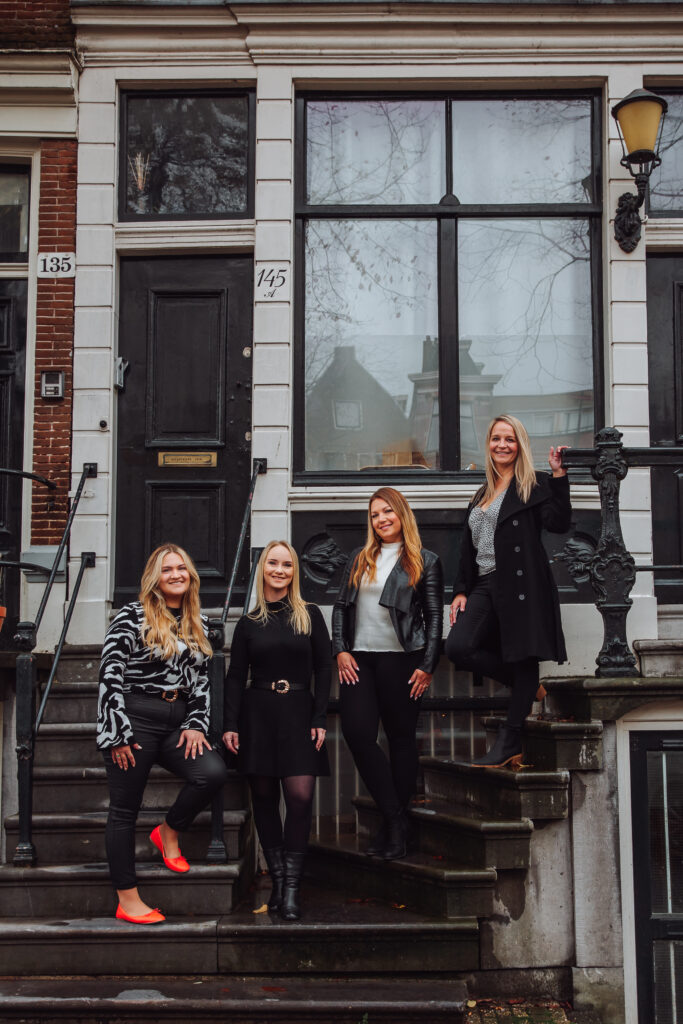 Four people standing on stone steps outside adjacent townhouses, dressed in black with one wearing red shoes.