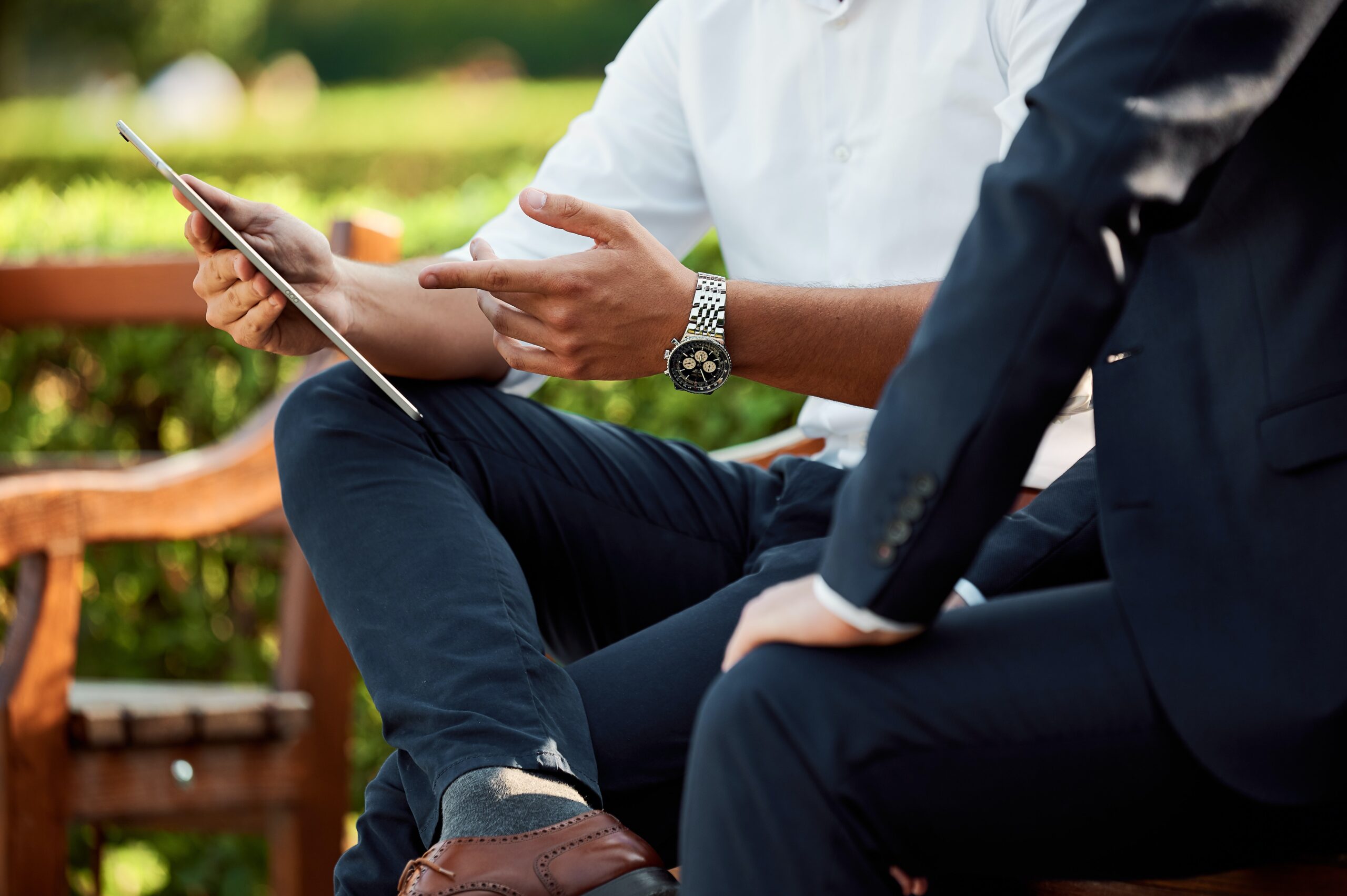 Services 6 Two individuals seated on wooden bench outdoors, one with tablet and the other in dark suit, engaged in discussion.