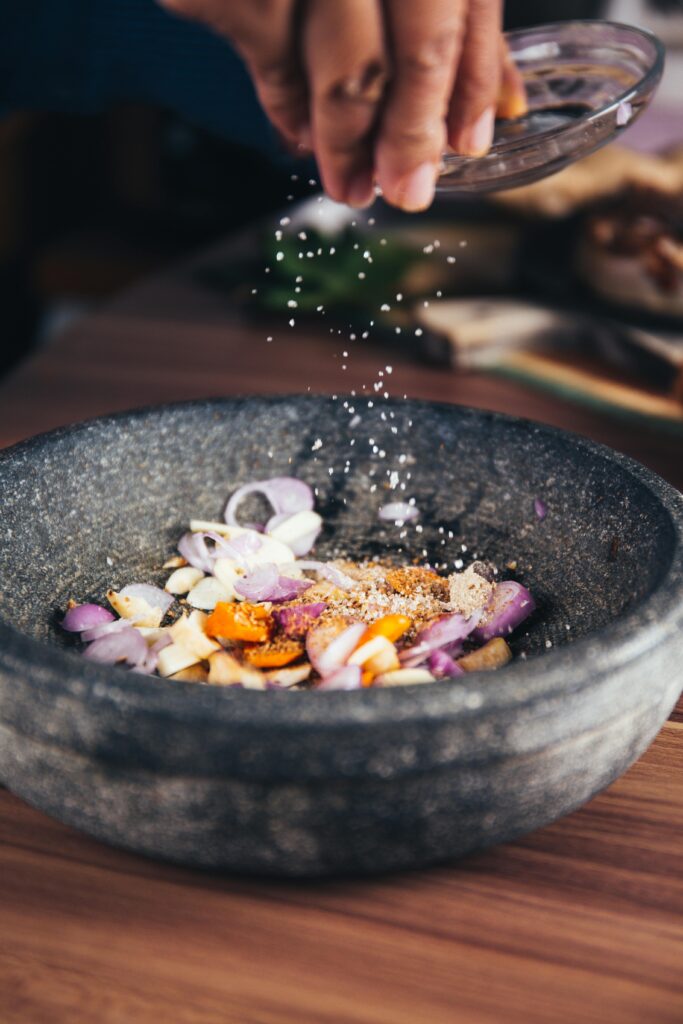 Hand sprinkling salt into a stone mortar with sliced shallots, garlic, and carrots on a wooden surface in a kitchen setting.