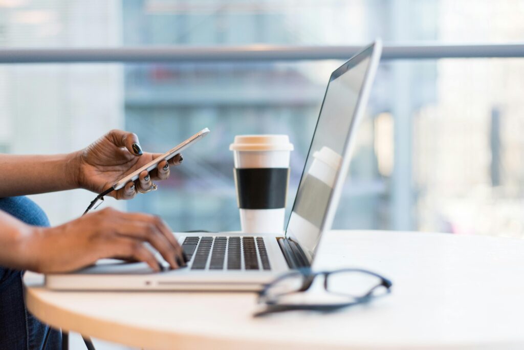 Close‑up of individual using laptop and smartphone at round table with eyeglasses and coffee cup.
