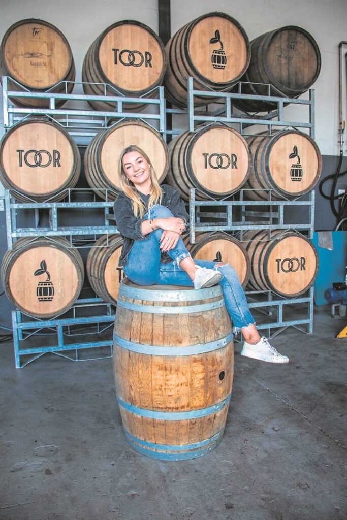 Person sitting on large wooden barrel inside distillery with stacked barrels labeled TOOR on metal racks.